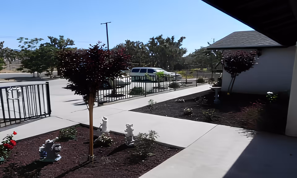 Outdoor view of a paved walkway with small landscaped garden areas featuring young trees and decorative statues. A white building with a sloped roof is partially visible on the right side, and a parked white vehicle is seen in the background near a black metal fence under a clear blue sky.