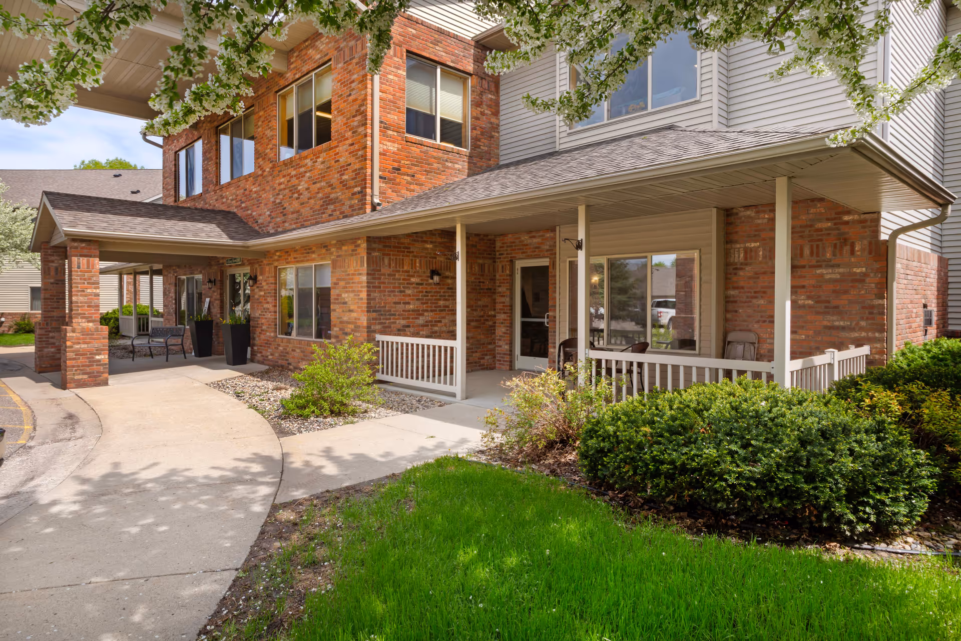 Exterior view of Mankato Lodge Senior Living showing a brick and siding building with a covered entrance, a curved concrete walkway, green bushes, and a grassy area under a tree with white blossoms.