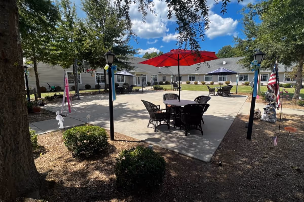 Outdoor patio area at MacGregor Downs Health Center with several round tables and chairs under large umbrellas. The patio is surrounded by a building with multiple windows and trees providing shade. There are lamp posts decorated with ribbons and an American flag visible on the right side.