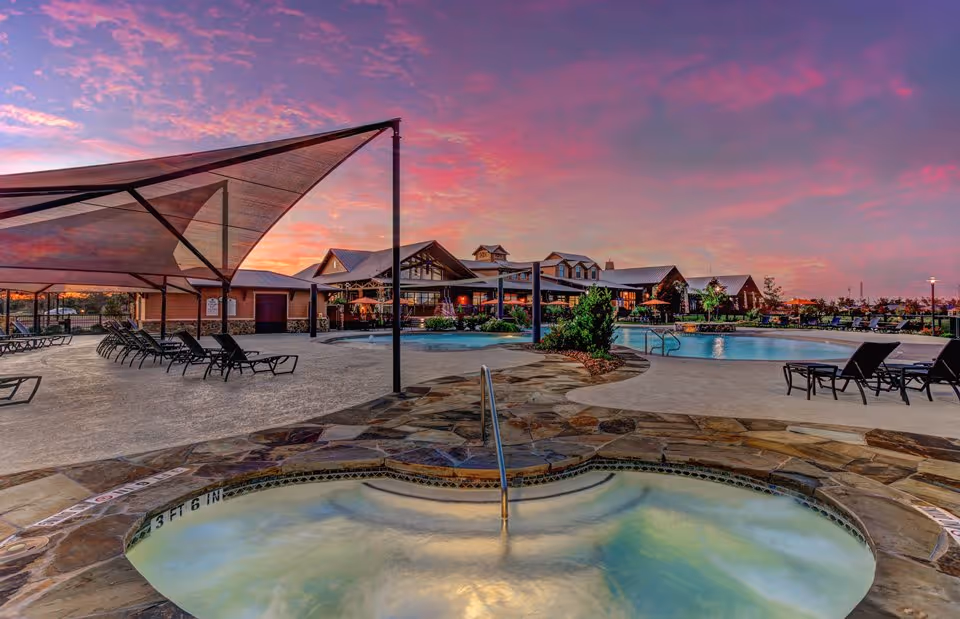 Resort-style outdoor pool and hot tub area with lounge chairs and a clubhouse under a colorful sunset.