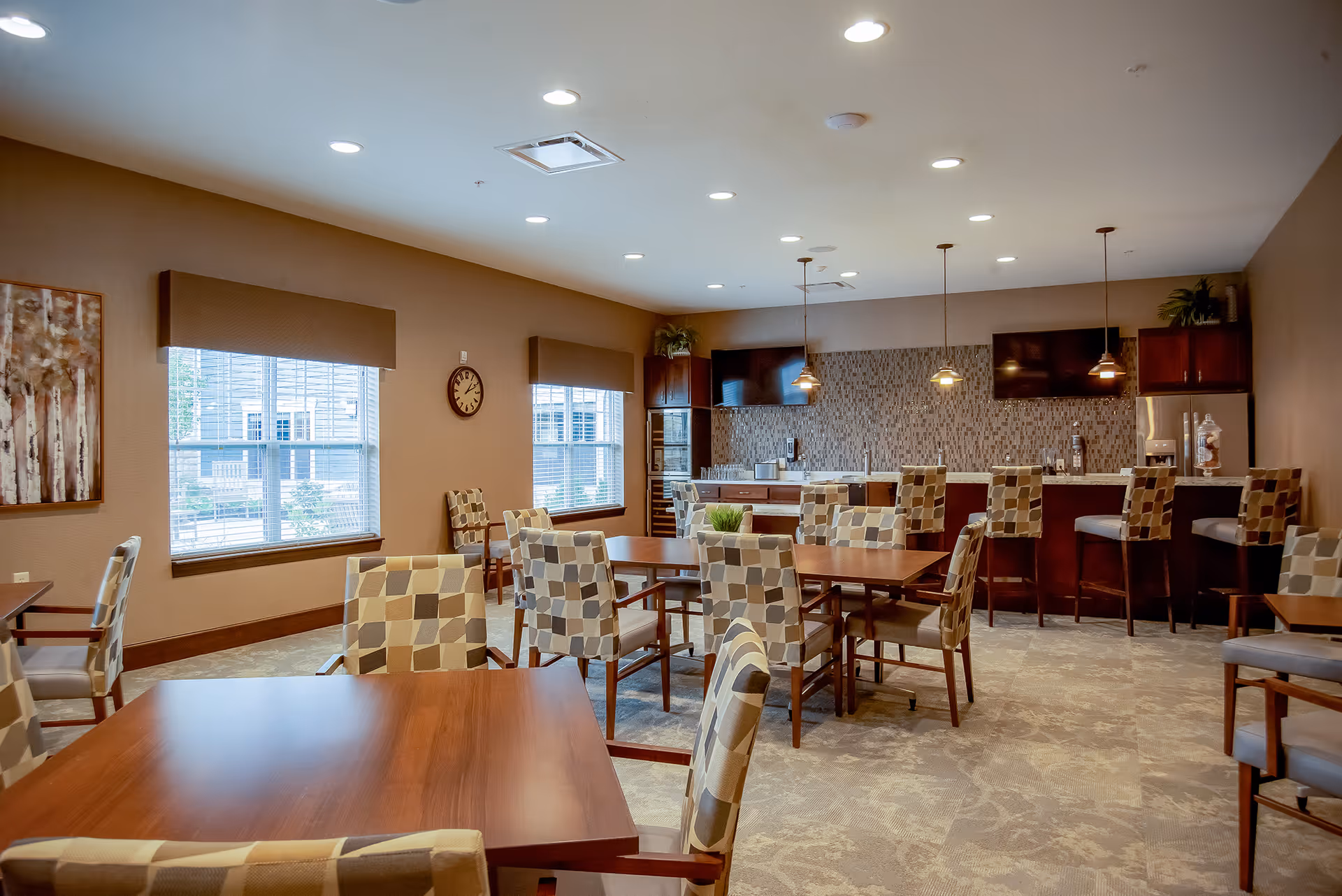A spacious dining area with multiple wooden tables and cushioned chairs featuring a geometric pattern. The room has beige walls, two large windows with brown valances, a wall clock, and a kitchen area in the background with a mosaic tile backsplash, pendant lights, and bar stools. The floor is carpeted with a subtle pattern.