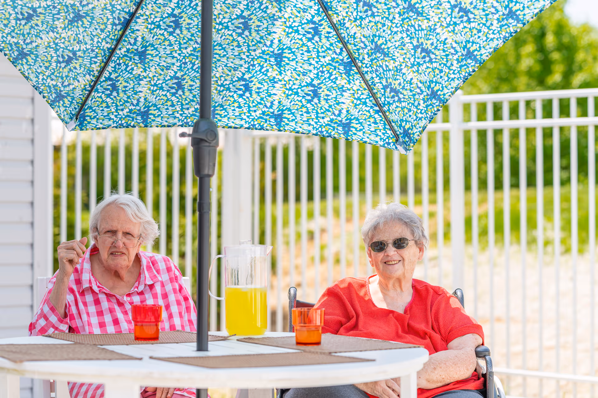 Two elderly women sitting outdoors at a round table under a blue patterned umbrella. One woman is wearing a red and white checkered shirt and glasses, while the other is wearing a red shirt and sunglasses. On the table, there is a pitcher of yellow drink and two orange cups. A white fence and greenery are visible in the background.