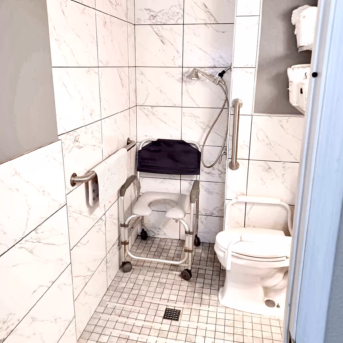 Accessible tiled bathroom with a commode chair, toilet with support rails, handheld shower and grab bars.