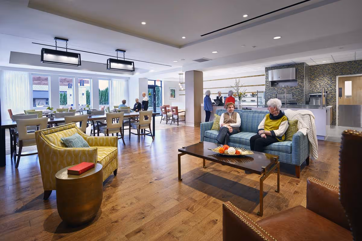 A spacious and well-lit common area in a senior living facility featuring a seating area with a blue sofa and two elderly women sitting and conversing. The room has wooden flooring, modern lighting fixtures, and several tables and chairs arranged for dining or socializing. In the background, other seniors and staff members are engaged in conversation near a kitchen area with stainless steel appliances and a decorative tiled wall.