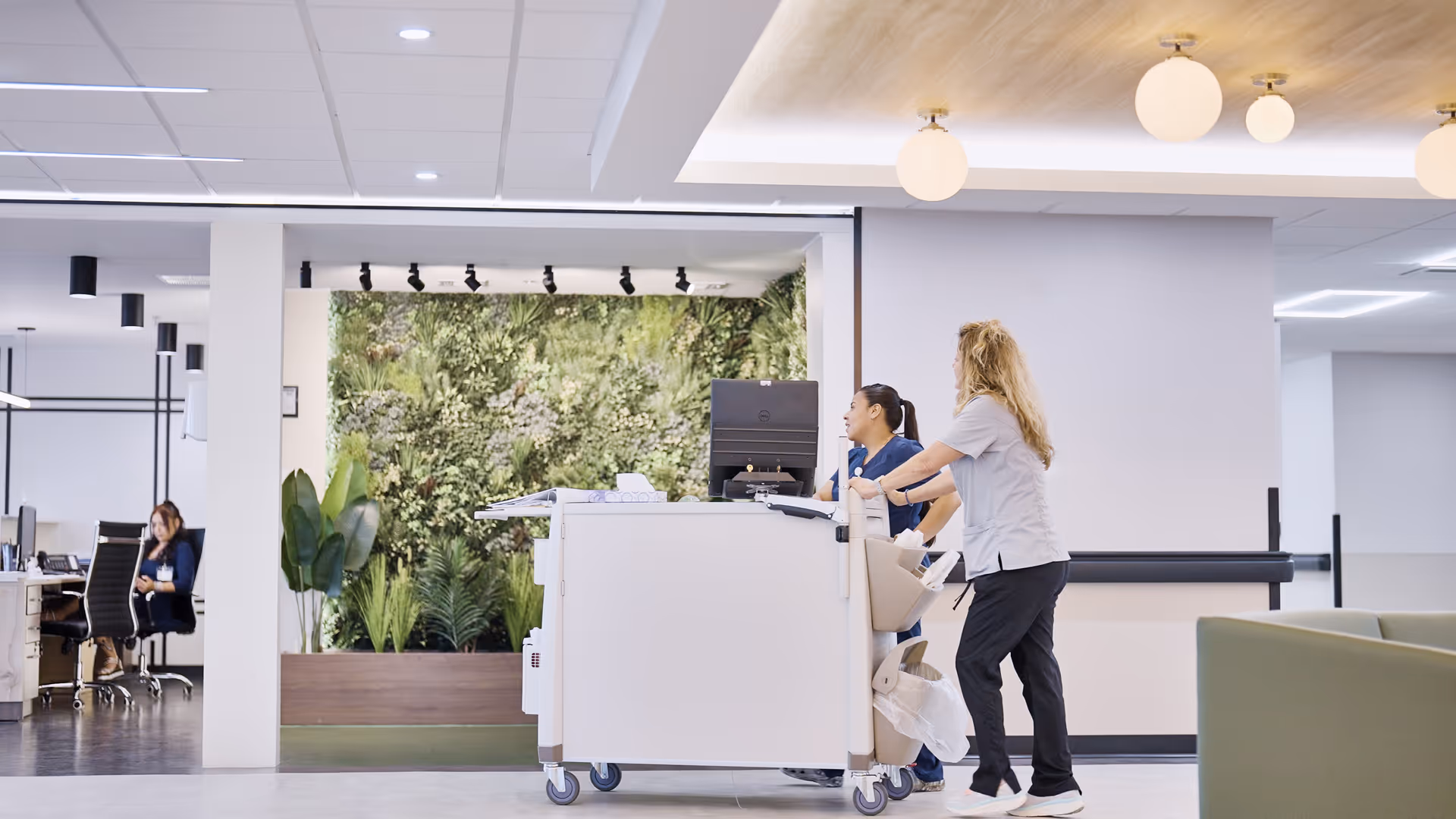 Two healthcare workers standing at a mobile workstation in a modern facility hallway with a green plant wall in the background and a woman sitting at a desk in an office area to the left.
