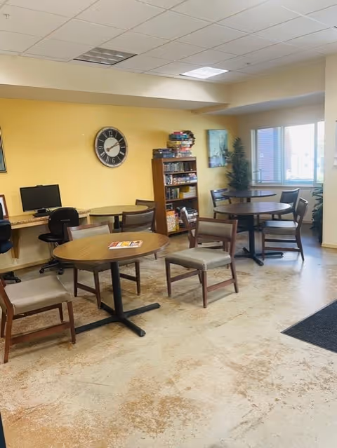 A common area with round wooden tables and chairs arranged around the room. There is a yellow wall with a large clock, a bookshelf filled with books and games, and a computer workstation with two chairs. A window lets in natural light, and there is a potted plant near the window.