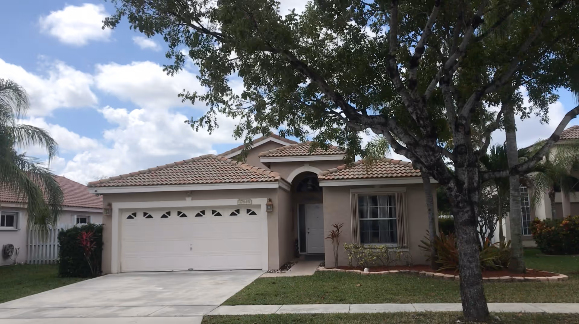 Front exterior view of a single-story house with a tiled roof, a two-car garage, a driveway, and a large tree in the front yard under a partly cloudy sky.