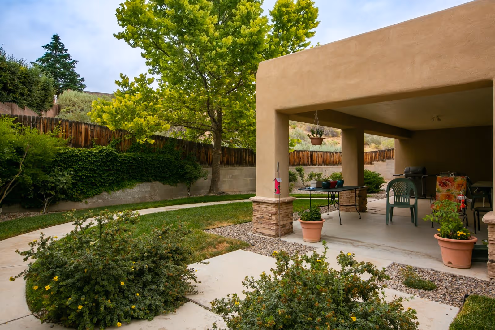 Outdoor patio area with a covered seating space featuring plastic chairs and a table with potted plants. Surrounding the patio is a garden with bushes, a tree, a wooden fence, and a concrete pathway.