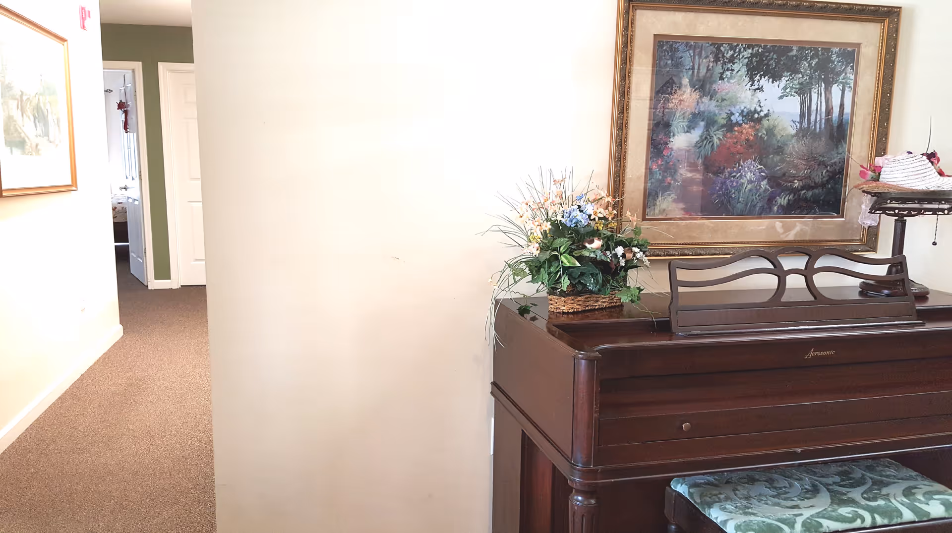 Interior view of a senior living facility hallway with a wooden piano on the right side. The piano has a floral arrangement on top and a framed painting of a garden scene hanging above it. There is a cushioned piano bench with a green patterned fabric in front of the piano. The hallway leads to a room with an open door, and another framed picture is visible on the left wall.