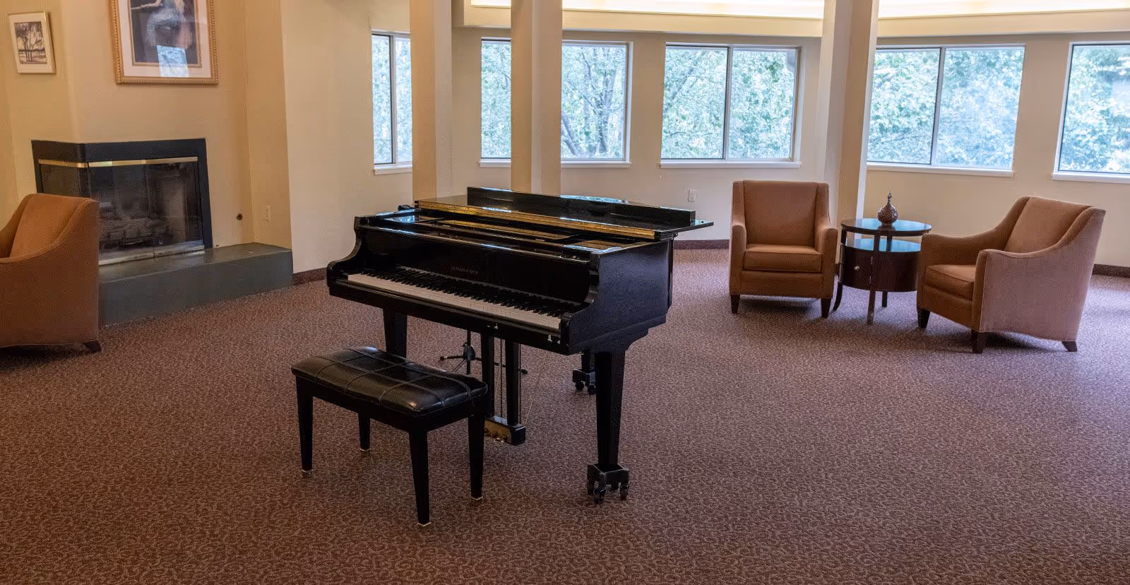 A spacious room with a black grand piano and matching bench in the center, surrounded by three brown armchairs and a round wooden side table. The room has large windows letting in natural light and a fireplace in the corner with framed artwork above it. The floor is carpeted with a patterned design.