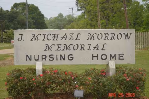 A stone sign reading I. Michael Morrow Memorial Nursing Home with blooming bushes.