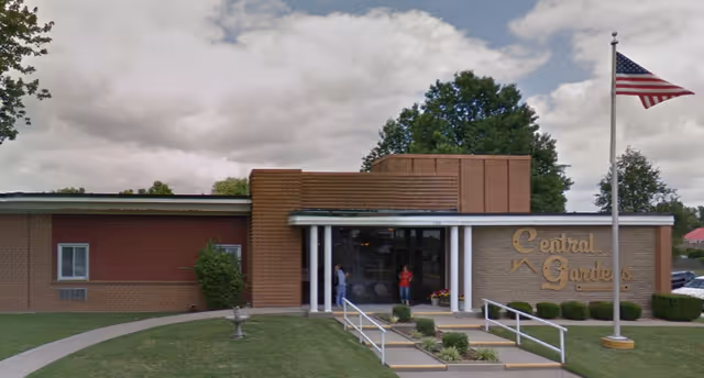 Front exterior view of a single-story brick building with a sign that reads 'Central Gardens'. There is a pathway leading to the entrance with handrails and small landscaped bushes. Two people are standing near the entrance. An American flag is flying on a flagpole to the right of the building. Trees and a partly cloudy sky are visible in the background.