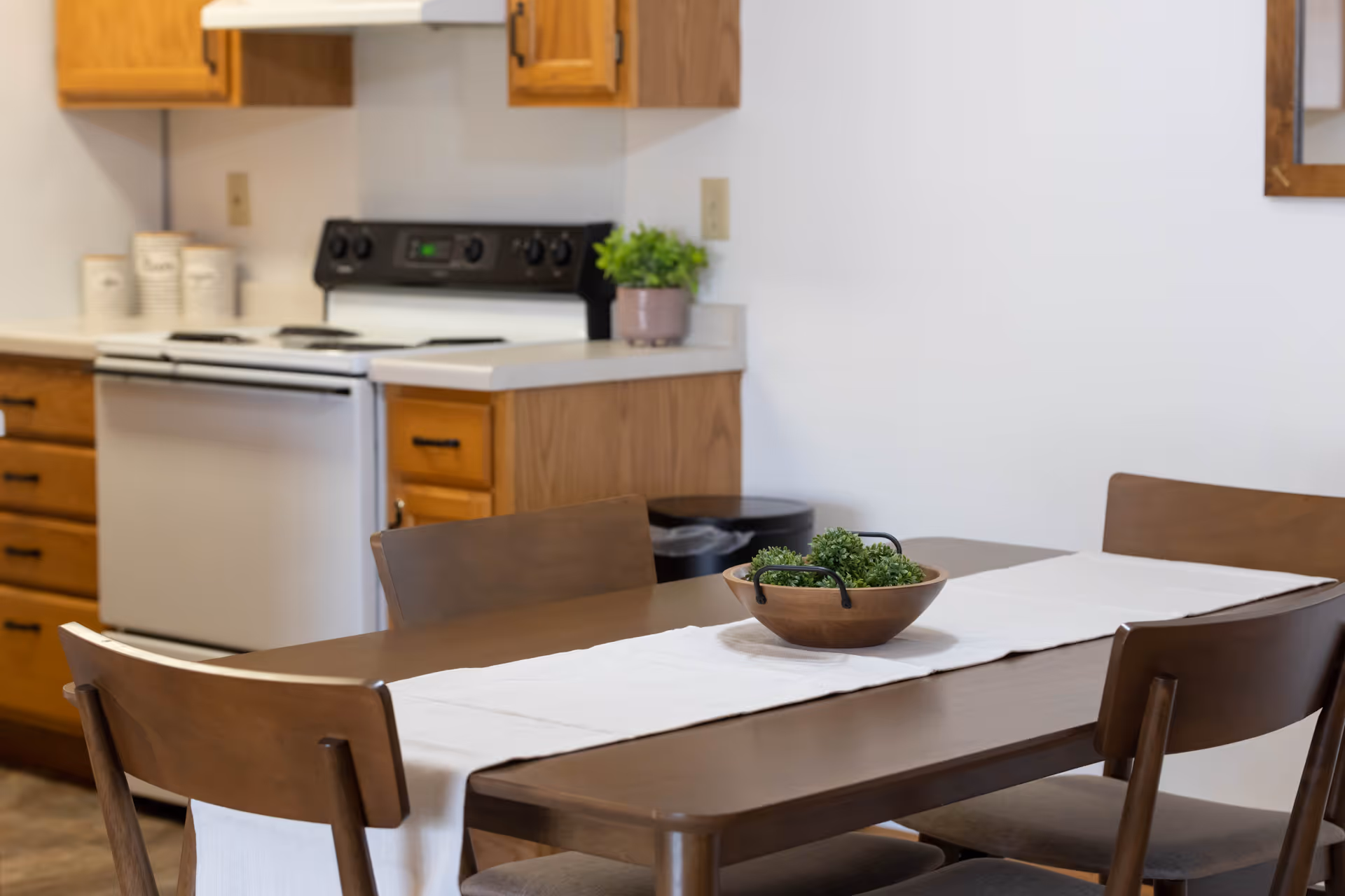 A kitchen and dining area with wooden cabinets, a white stove, and a wooden dining table with four chairs. The table has a white runner and a wooden bowl with green decorative plants. There is a small potted plant on the kitchen counter.