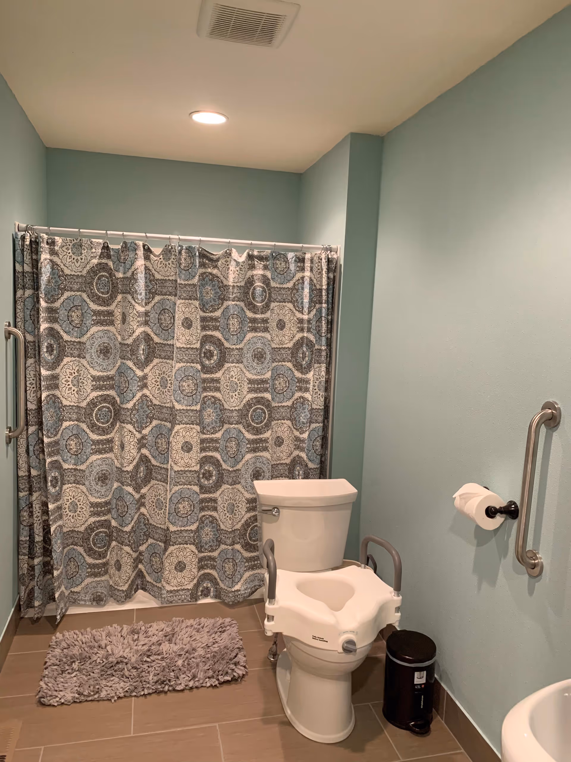 A bathroom with light blue walls and a tiled floor. There is a toilet with a raised seat and armrests, a patterned shower curtain with blue and gray circular designs, a gray shaggy bath mat, a metal grab bar on the wall next to the toilet with a toilet paper holder, and a small black trash can on the floor.