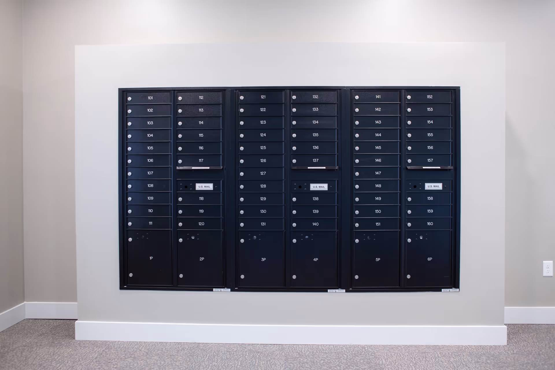 A wall-mounted set of black mailboxes with numbered compartments arranged in six columns, each column containing multiple small locked boxes and a few larger locked boxes at the bottom. The mailboxes are installed on a light-colored wall with a carpeted floor below.