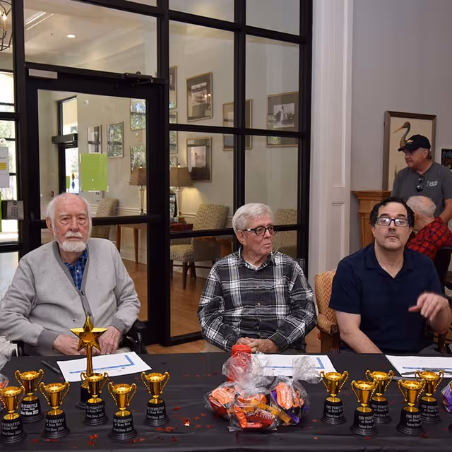 Three elderly men sitting at a table with small trophies and wrapped candy in front of them inside a room with glass doors and framed pictures on the walls. Two other men are visible in the background near a fireplace.