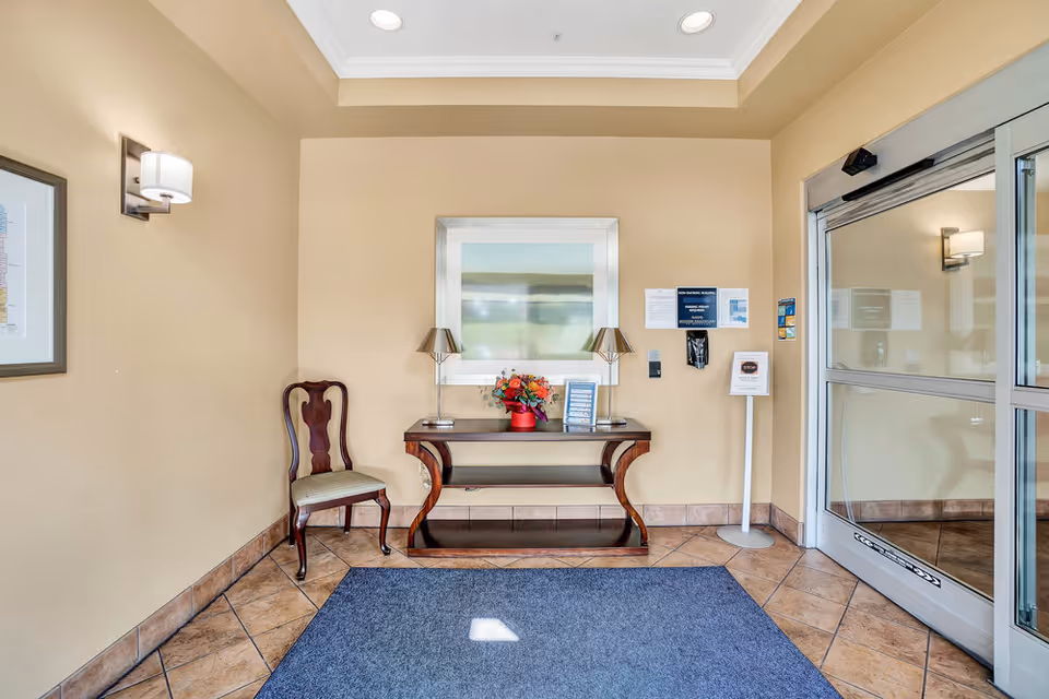 Entrance area of a facility with a glass automatic sliding door on the right, a wooden chair with a cushioned seat on the left, and a wooden console table in the center holding two lamps and a vase with flowers. A framed abstract artwork hangs above the table, and informational signs are posted on the wall near the door. The floor is tiled with a blue rug in front of the door.