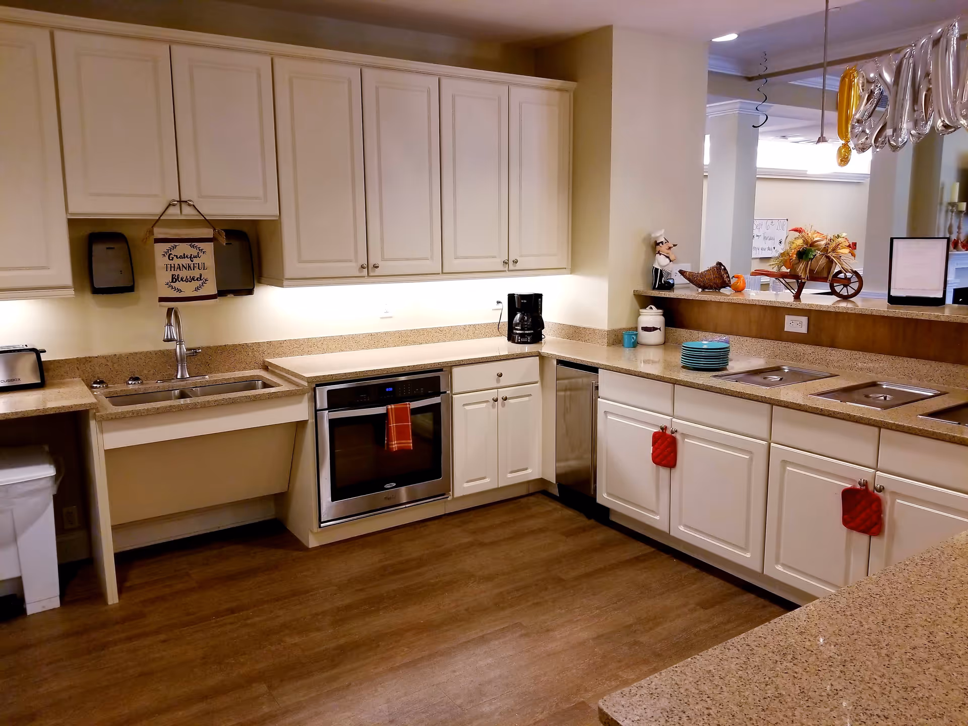 Bright communal kitchen with white cabinets, stainless appliances, sinks, and a countertop serving area decorated with plates and fall decor.