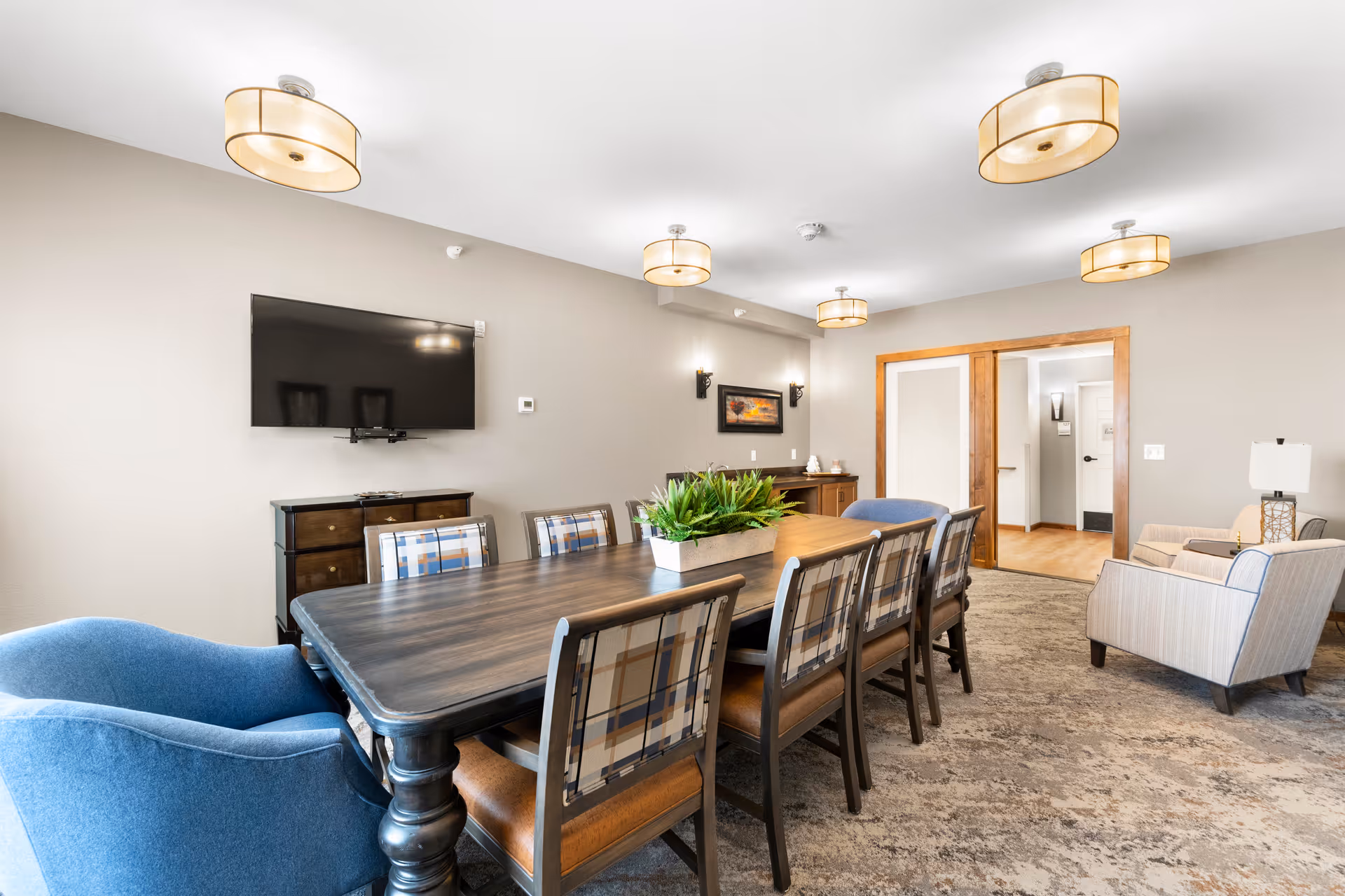 A well-lit senior living facility common room with a long wooden table surrounded by eight chairs, including two blue armchairs at the ends. The room features beige walls, a mounted flat-screen TV, a sideboard with decorative items, and modern ceiling lights. There is a doorway leading to another area of the facility.