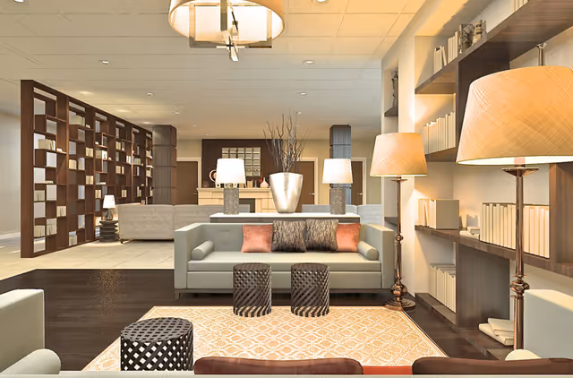 A modern and spacious living room area with a light gray sofa adorned with decorative pillows, two small round stools in front, and a patterned rug underneath. The room features warm lighting from floor lamps and a ceiling light fixture. There are built-in shelves filled with books on the right wall and a large wooden partition with open shelving on the left. In the background, additional seating and a decorative vase with branches are visible.