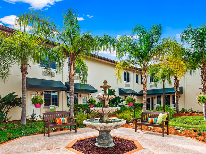 Outdoor courtyard area at The Windsor of Venice featuring a three-tier stone fountain in the center, surrounded by palm trees, green grass, colorful hanging flower pots, and two wooden benches with colorful cushions under a bright blue sky.