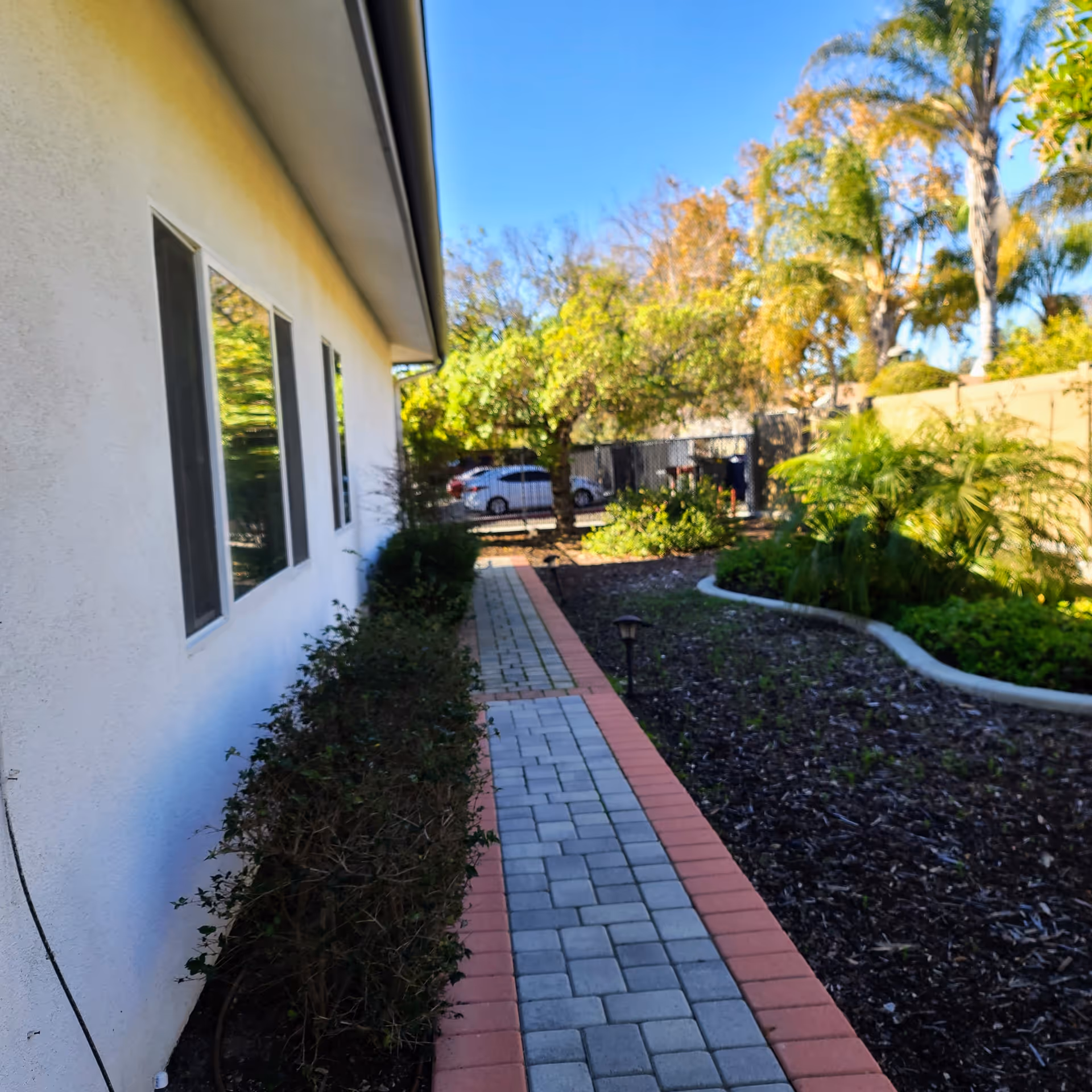 Paved brick pathway running beside a white building with landscaped beds and trees leading to a fenced area with parked cars.