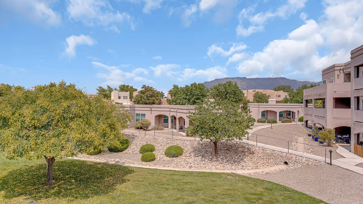 Outdoor view of Bear Canyon Estates senior living facility showing a landscaped courtyard with trees, bushes, gravel paths, and beige buildings under a partly cloudy blue sky with mountains in the background.
