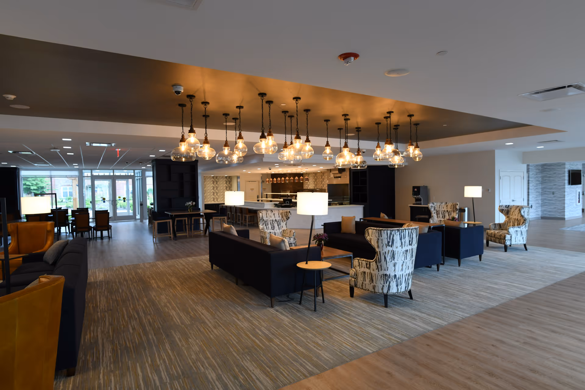 Open contemporary common lounge with sofas, patterned armchairs, round pendant lights, and a kitchenette/bar area.