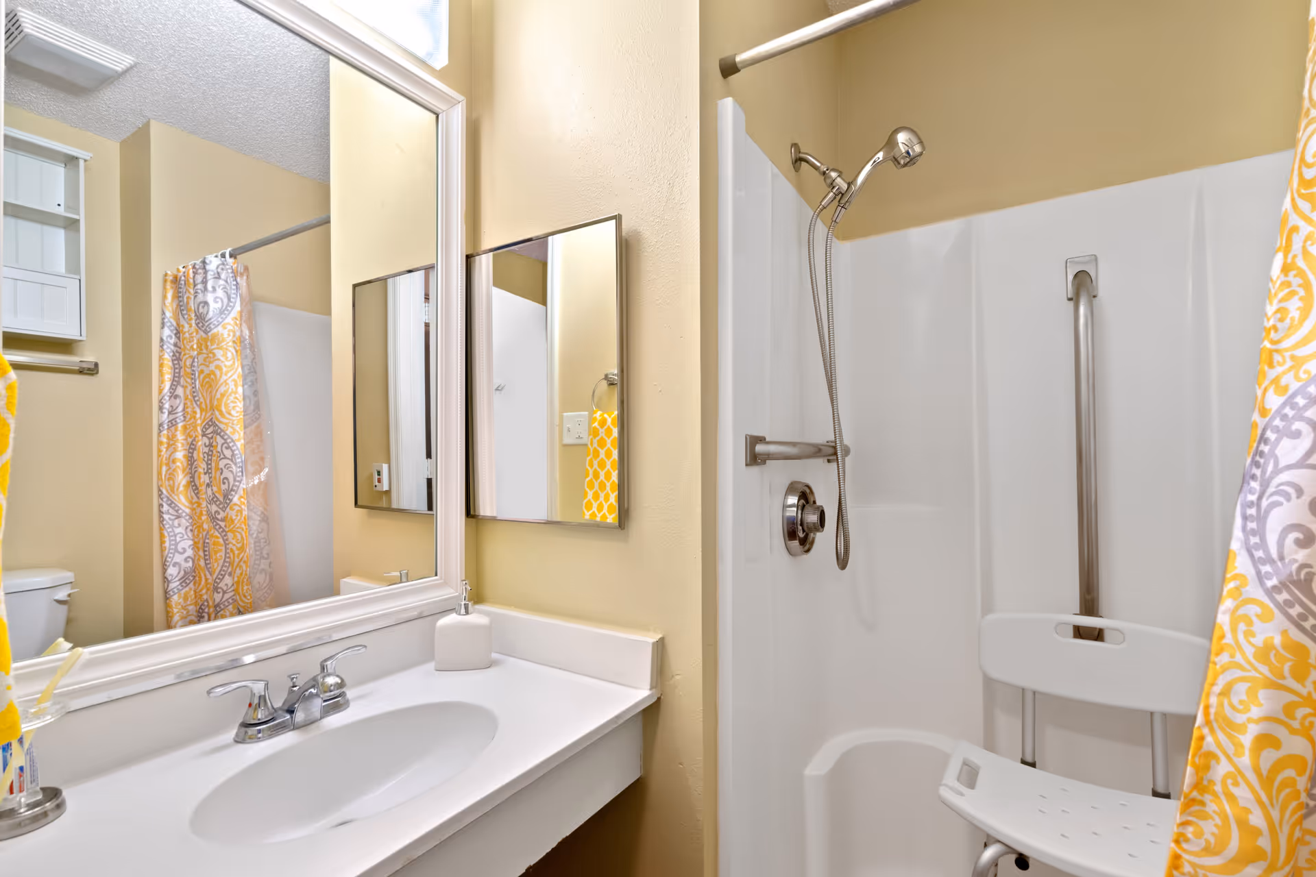 Bathroom with a white sink and countertop, a large mirror above the sink, a shower area with a handheld showerhead, a grab bar, and a white shower chair. The shower curtain has a yellow and gray decorative pattern. The walls are painted light beige.