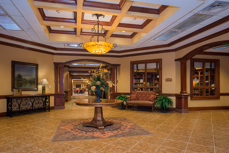 Spacious interior common area with a round wooden table holding a large floral arrangement in the center on a decorative rug. The room features tiled flooring, warm beige walls with wooden trim, a decorative ceiling light fixture, a cushioned bench with plants on either side, a painting on the wall, and a view into another room with tables and chairs.