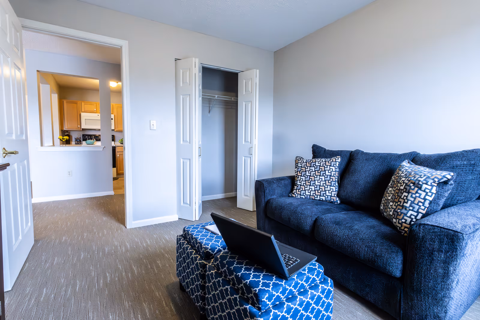 Interior view of a living room area in an apartment with a dark blue sofa adorned with patterned pillows. In front of the sofa is a blue patterned ottoman with a laptop on top. The room has beige carpet flooring and light gray walls. An open closet with white bi-fold doors is visible, and through an open doorway, a kitchen area with wooden cabinets and a microwave can be seen.