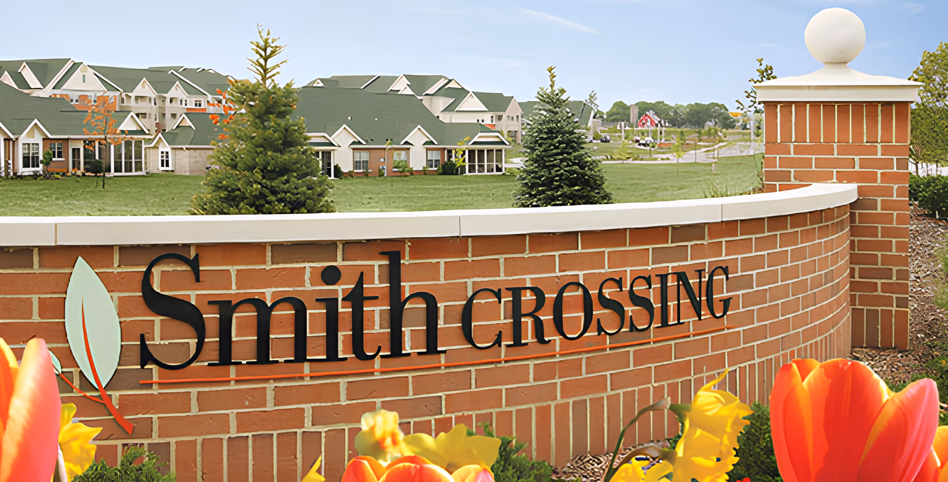 Brick entrance sign reading "Smith Crossing" with flowers, lawn, and facility buildings behind it.