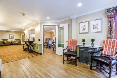 Interior view of a senior living facility hallway with wooden flooring and beige walls. Two wooden chairs with red plaid cushions flank a small black cabinet topped with three small potted plants. Two framed floral artworks hang above the cabinet. In the background, an open door leads to a room with additional seating. The hallway extends to the left with more chairs and a decorative table along the wall.