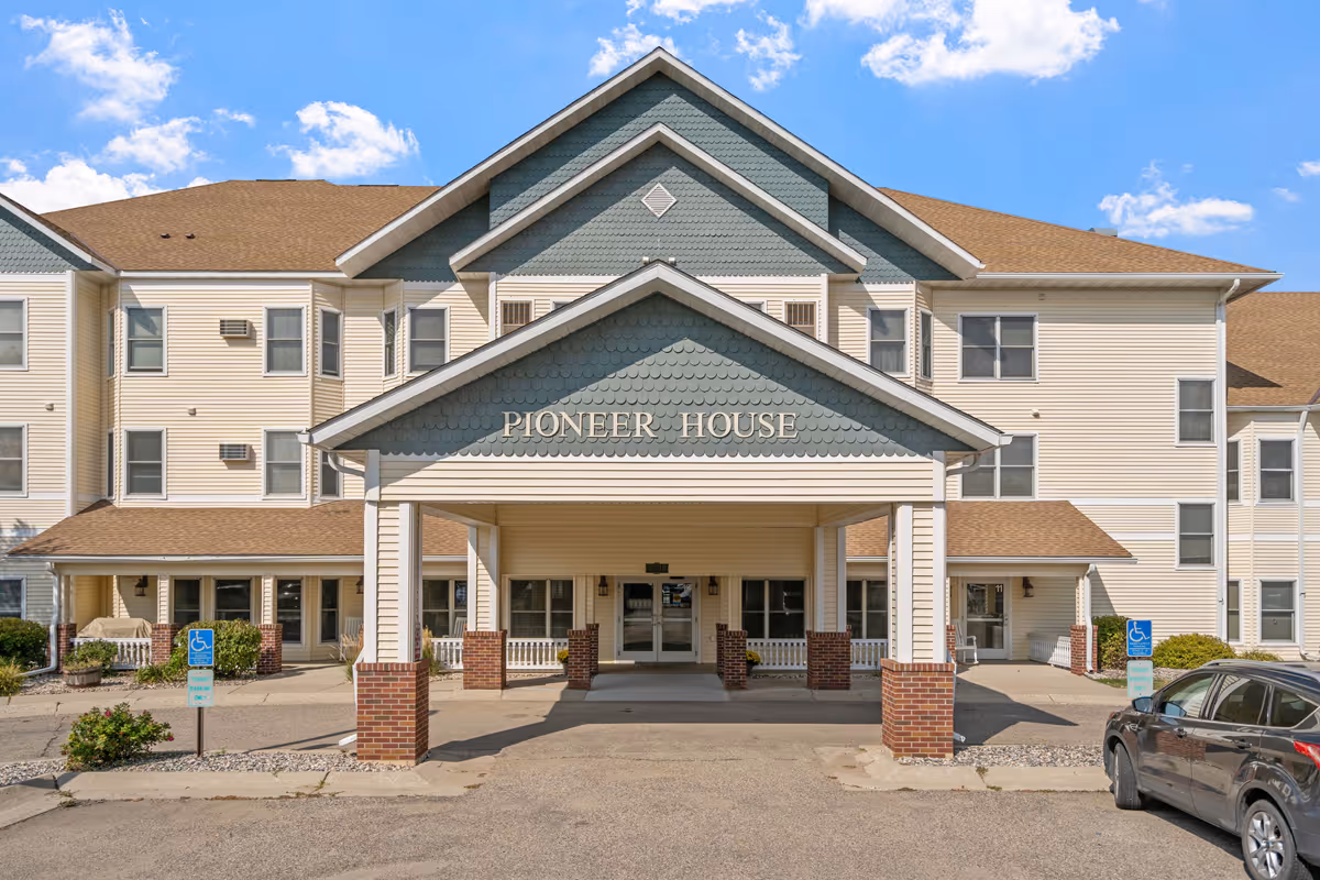 Front exterior view of a three-story senior living community building named Pioneer House with a covered entrance supported by brick and white columns. There are handicap parking signs and a parked car visible in front of the building under a partly cloudy blue sky.