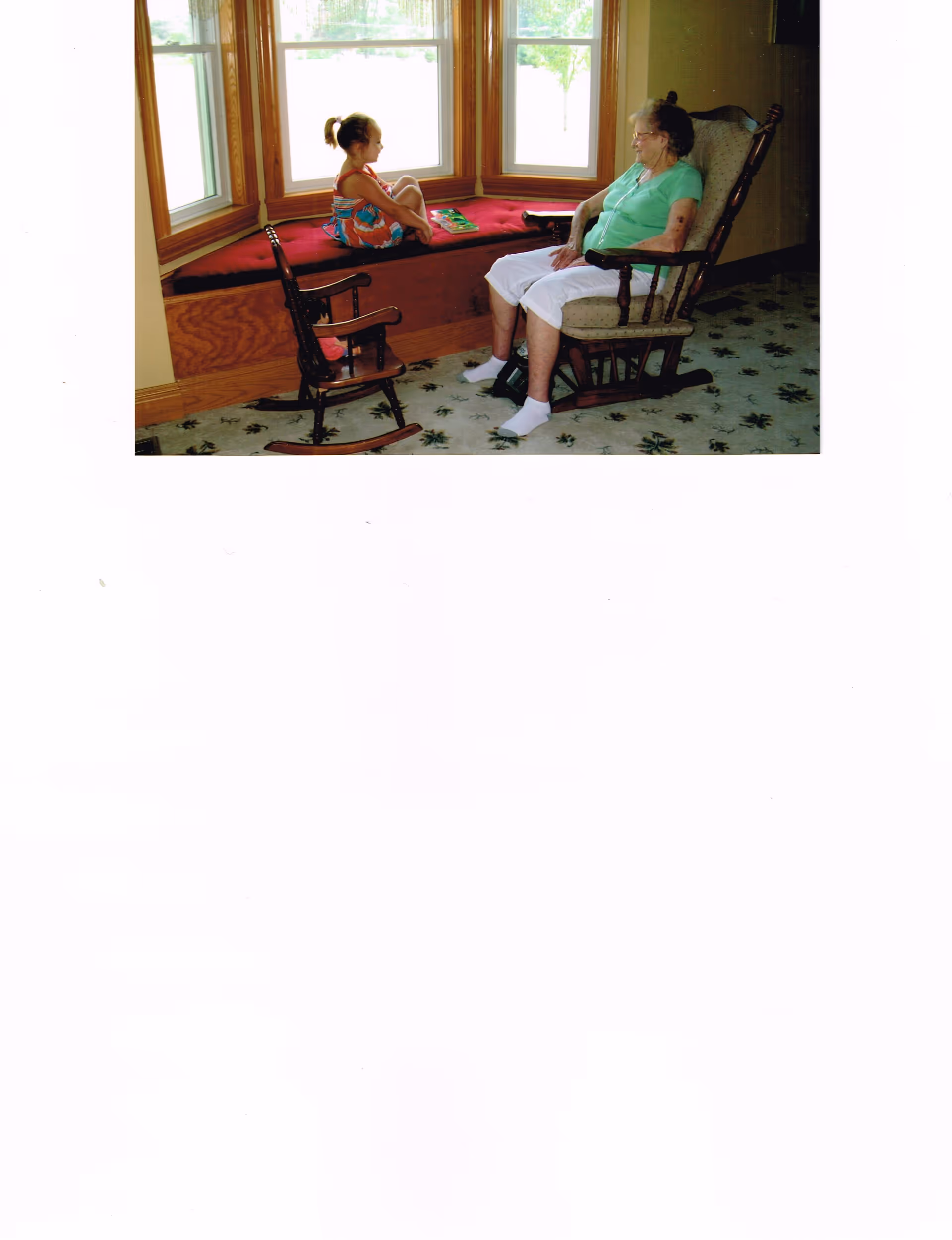 An elderly woman sitting in a cushioned rocking chair facing a young girl who is sitting cross-legged on a window seat with a red cushion. The girl is wearing a colorful dress and has a book beside her. There is also a small wooden rocking chair between them. The room has large windows with wooden frames and a carpeted floor with a floral pattern.