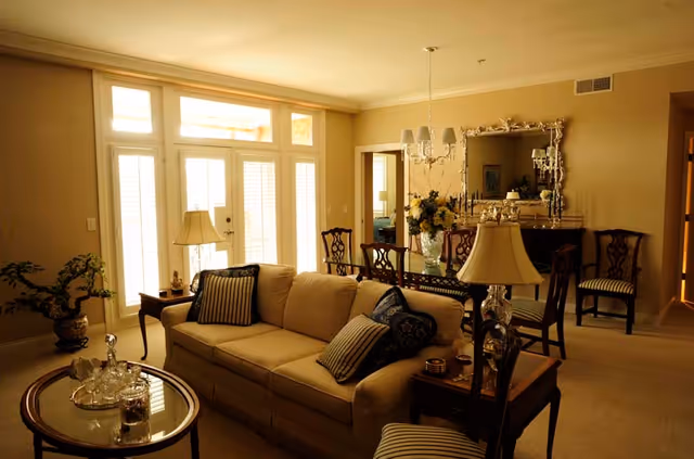 Sunlit living room with a beige sofa, striped pillows, a glass-top coffee table and a dining area with chandelier in the background.