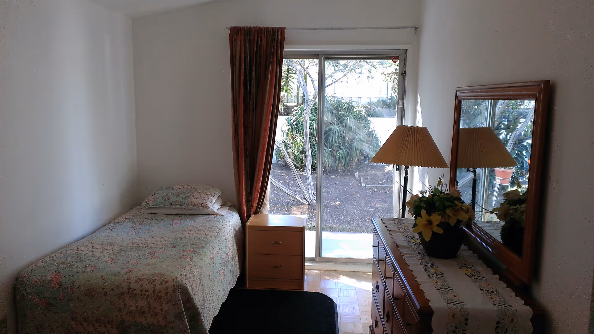 Small bedroom with a single bed, nightstand, dresser with lamp and mirror, and sliding glass doors opening to a garden.