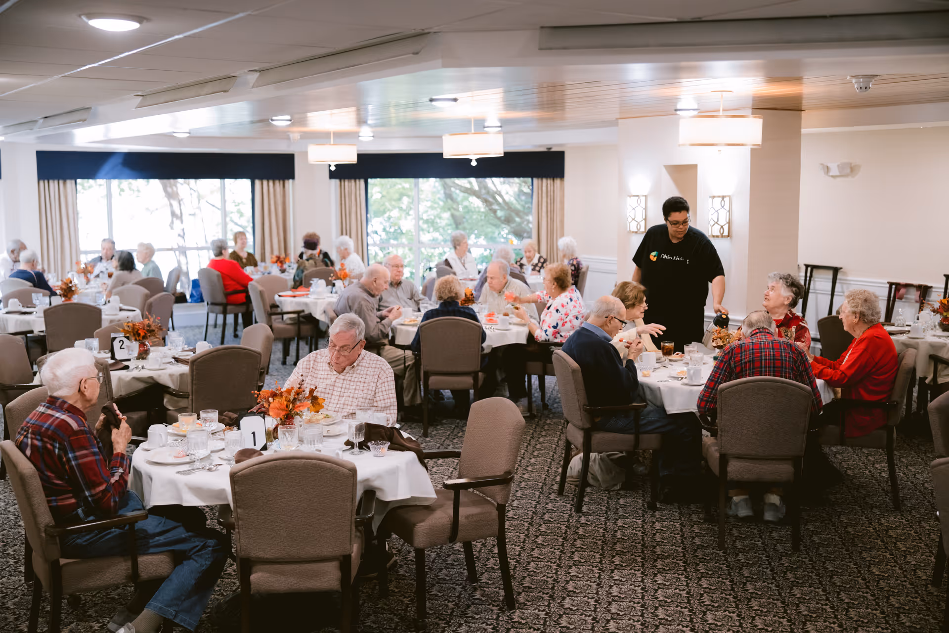 A dining room in a senior living facility with elderly residents seated at round tables covered with white tablecloths. A staff member is serving drinks to one of the tables. The room has large windows with curtains, patterned carpet, and ceiling lights.
