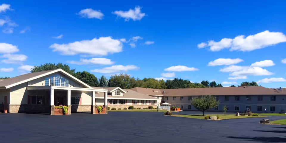 Two-story senior living building with a covered entrance, landscaped grounds, and a large paved driveway under a blue sky.