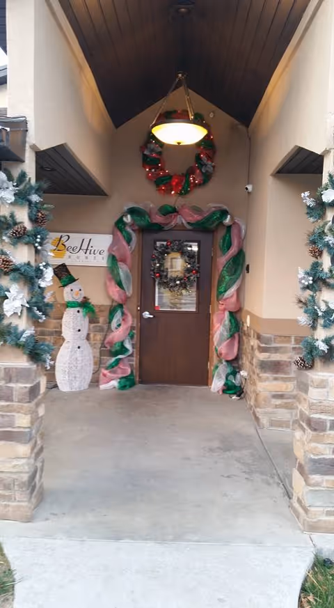 Entrance to Beehive Homes of Payson decorated for Christmas with a wreath on the door, a large candy cane garland around the door frame, a hanging wreath above the door, and a snowman decoration to the left. The entrance has stone pillars adorned with garlands and pine cones.