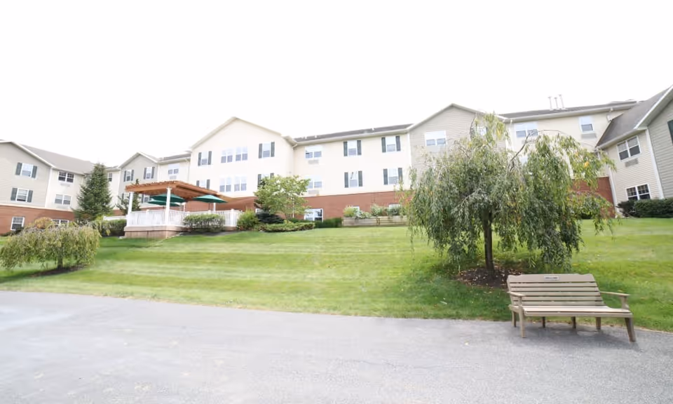 Exterior view of a multi-story senior living facility with beige siding and brick accents. The building is surrounded by a well-maintained lawn with small trees and shrubs. There is a wooden bench and a tree in the foreground, and a covered patio area with green umbrellas is visible on the left side of the building.