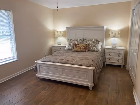 A neatly made bed with a white wooden frame and headboard, flanked by two matching white nightstands each with a lamp. The bed has a gray comforter and decorative pillows. The room has beige walls, wooden flooring, a window with blinds on the left, and a closed door on the right.