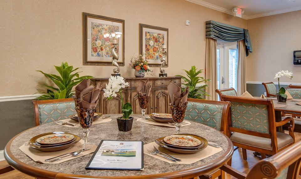A dining room in South Breeze Senior Living featuring a round table set with plates, napkins, utensils, and glasses. The table has a small potted orchid in the center and a menu labeled 'South Breeze'. The room has wooden chairs with patterned upholstery, plants, framed floral artwork on the wall, and a sideboard with decorative items. There is a door with curtains leading outside.