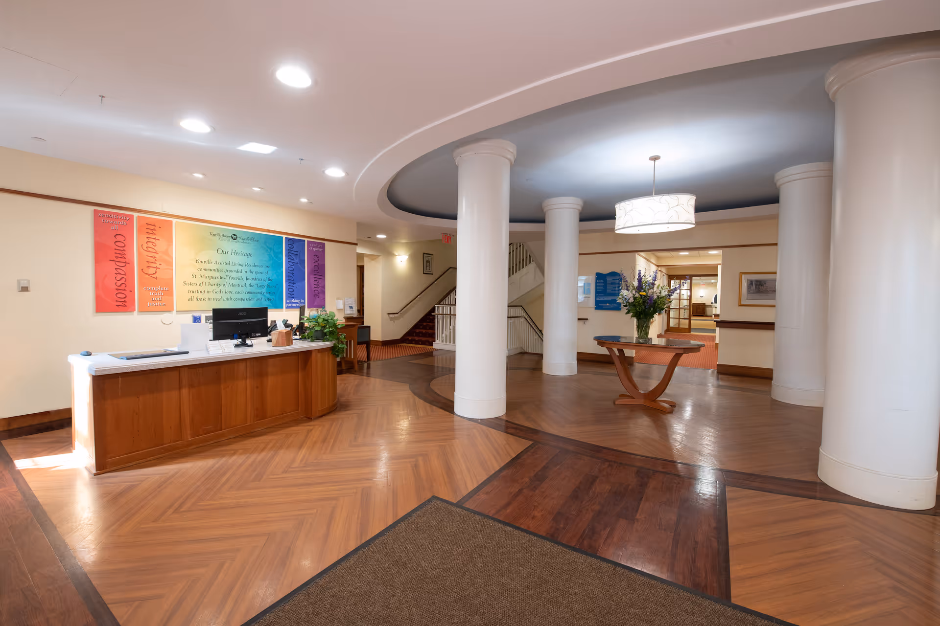 Reception area of Youville Place featuring a wooden reception desk with a computer and plants, a round wooden table with a flower arrangement, large white columns, and colorful banners on the wall displaying values such as compassion, integrity, collaboration, and excellence.