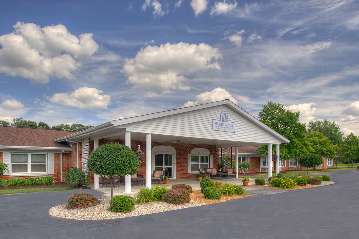 Front entrance of a single-story brick senior living facility with a covered porte-cochère, seating, and landscaped beds under a cloudy sky.