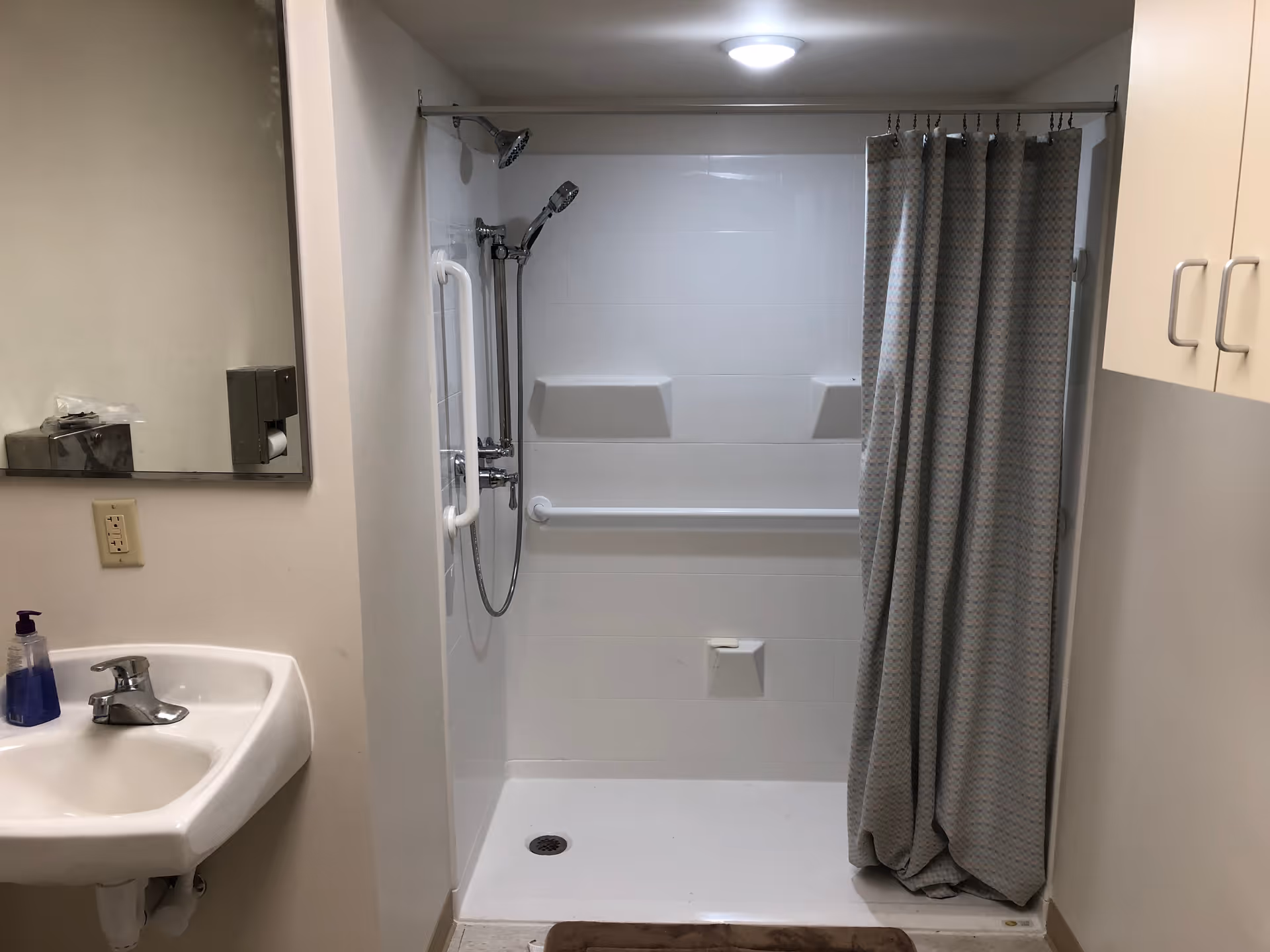 A bathroom with a white sink and faucet on the left, a large mirror above the sink, and a walk-in shower with a grab bar and a shower curtain on the right. There are beige cabinets on the far right wall and a brown bath mat on the floor in front of the shower.