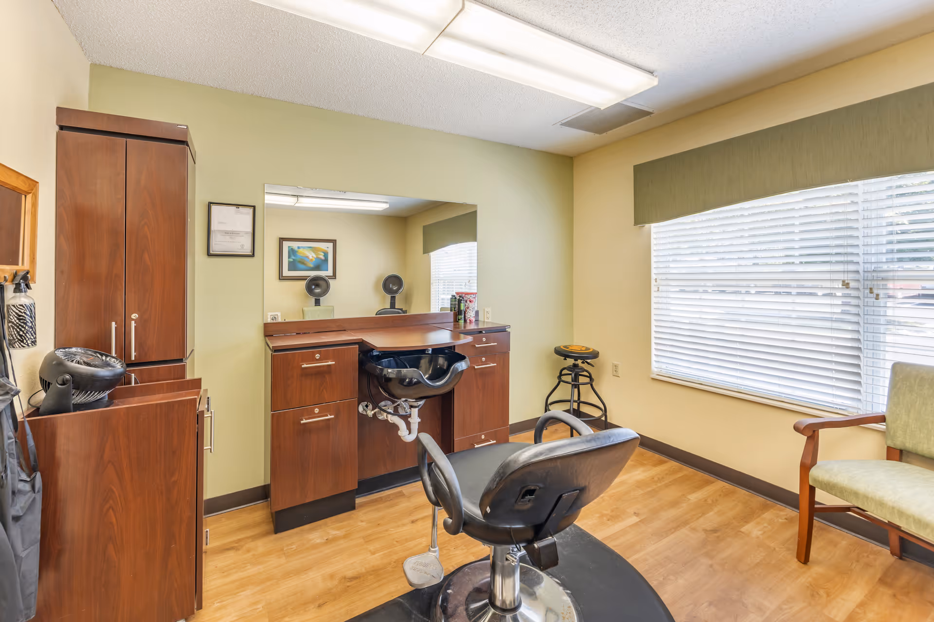 A small hair salon room with a black salon chair in front of a wooden counter with a built-in black sink. There is a large mirror above the counter, a wooden cabinet to the left, a stool near the window with blinds, and a green cushioned chair to the right. The room has light yellow walls and wood flooring.