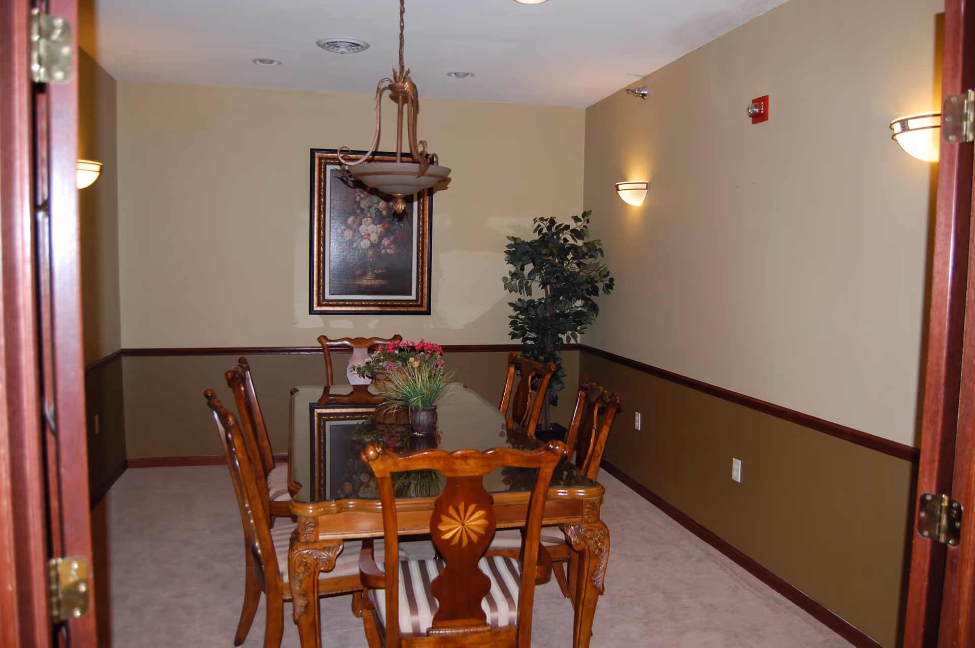 A dining room with a wooden table and six matching chairs with striped cushions. The table has a glass top and a floral centerpiece. The walls are painted beige with a darker brown wainscoting. There is a framed floral painting on the far wall and a potted plant in the corner. The room is lit by wall sconces and a hanging light fixture above the table.