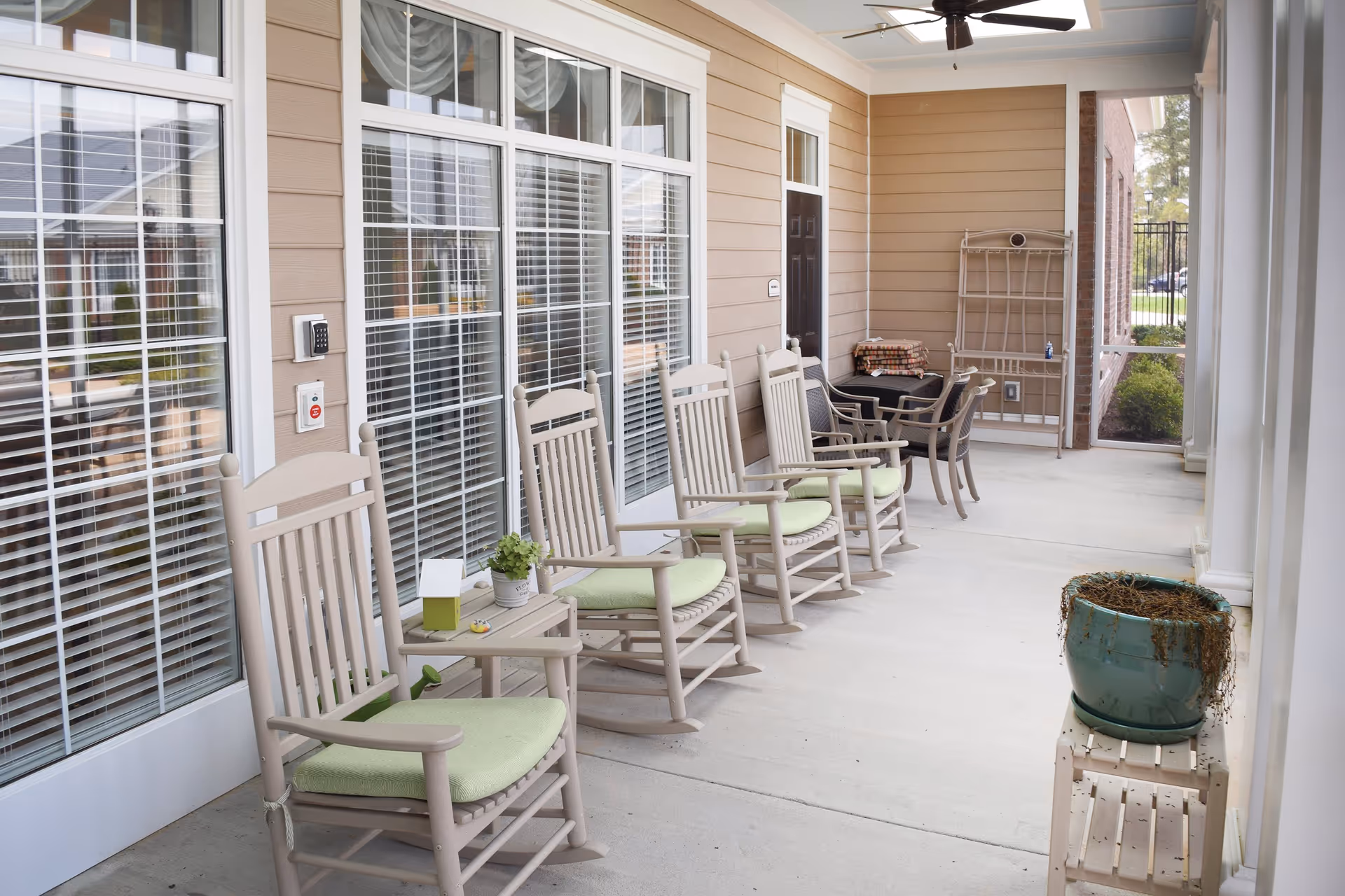 A covered outdoor patio area at Spring Arbor Cottage of Salisbury featuring a row of beige rocking chairs with light green cushions, a small side table with a plant and tissue box, a ceiling fan, and a large potted plant on a small stand. The patio is adjacent to a building with large windows and a door.