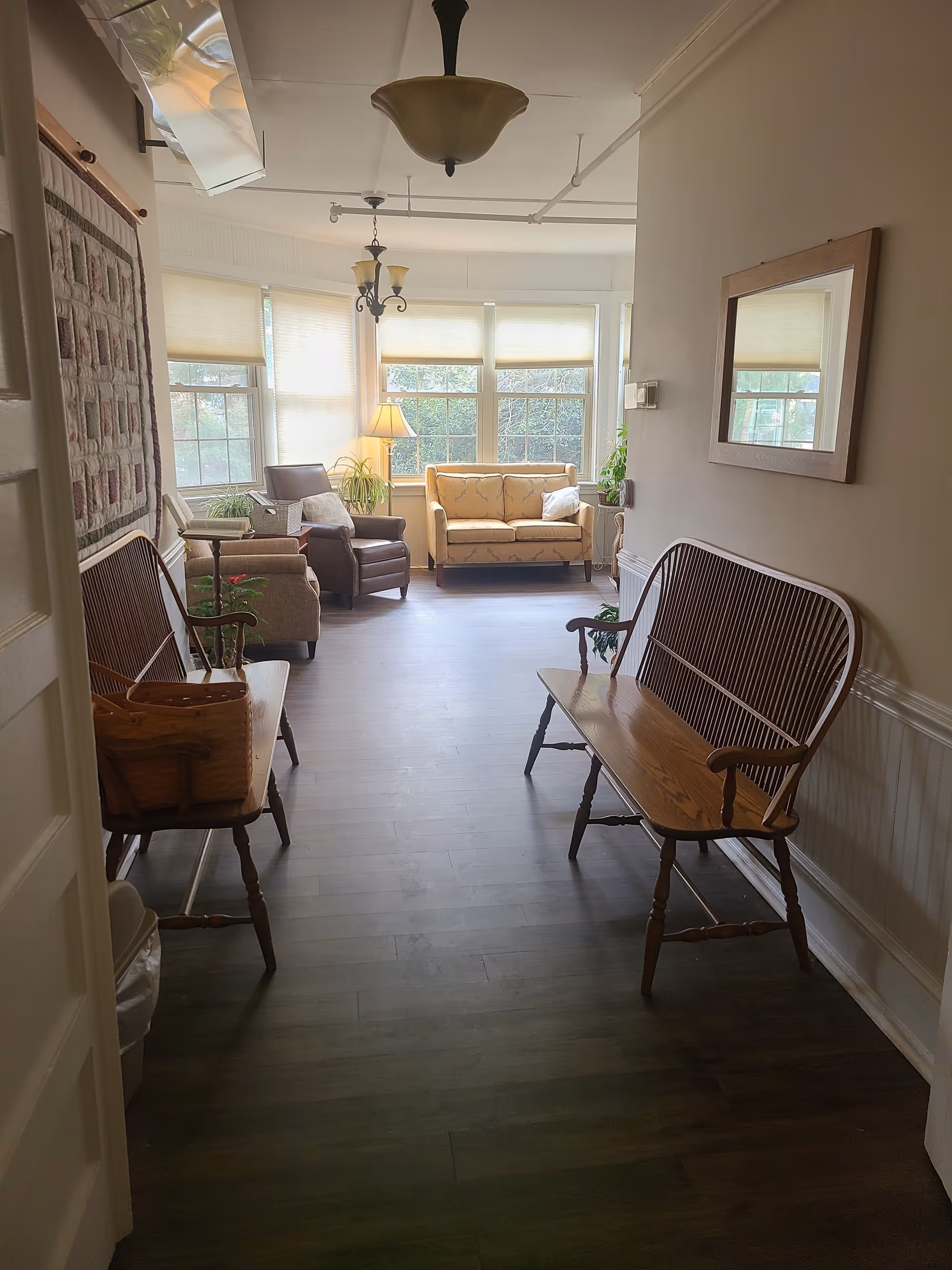 A bright and cozy sitting area with large windows letting in natural light. The room features a wooden bench on each side of the hallway, a beige loveseat, two armchairs, a floor lamp, and several potted plants. The walls are light-colored with a framed mirror on the right wall and a quilt hanging on the left wall.